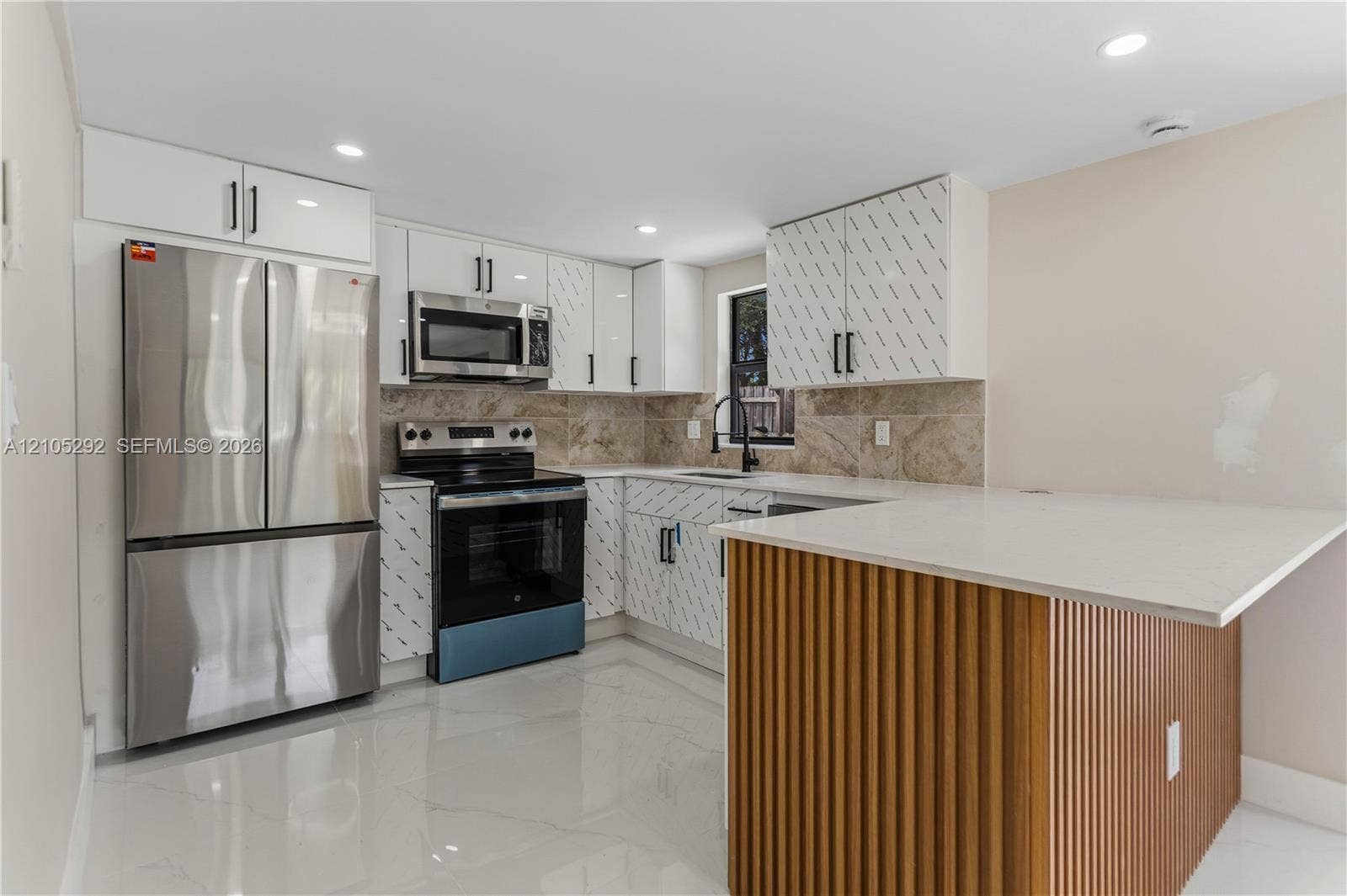 Modern kitchen featuring stainless steel appliances, white cabinetry with patterned accents, stone backsplash, and an island with a slatted wood-finish base and light-toned countertop