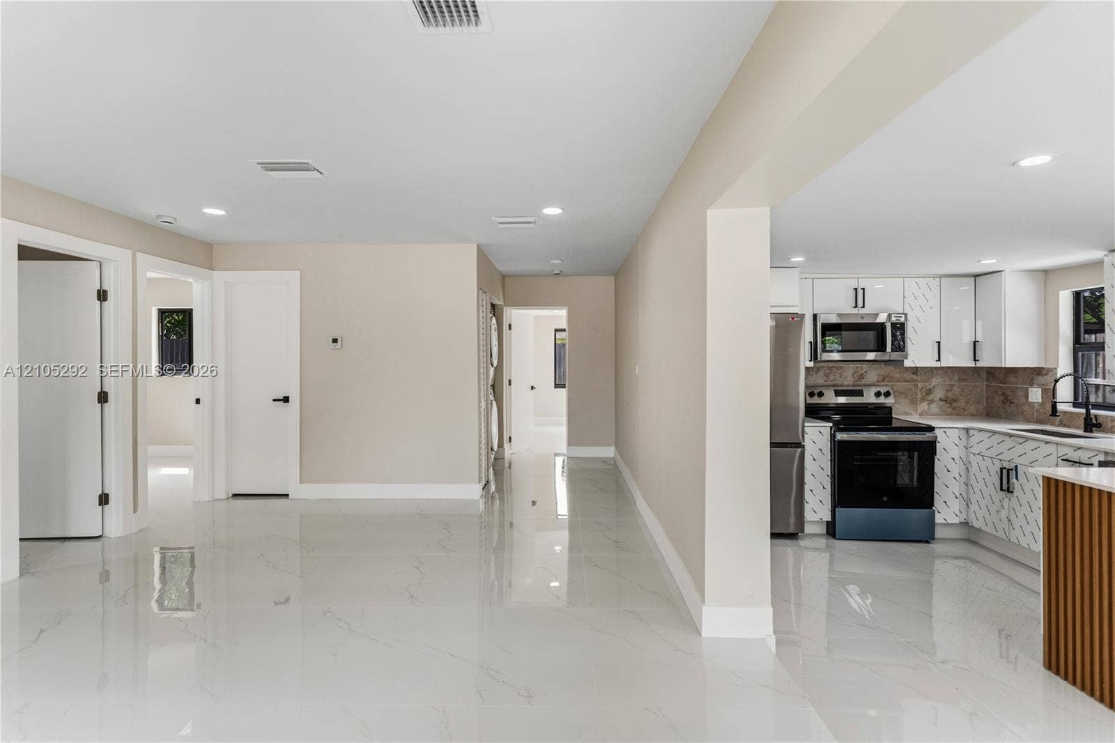 Glossy white tile flooring, recessed ceiling lighting, light-colored wall paint, white interior doors with dark hardware, and a partial view of a kitchen with white cabinetry