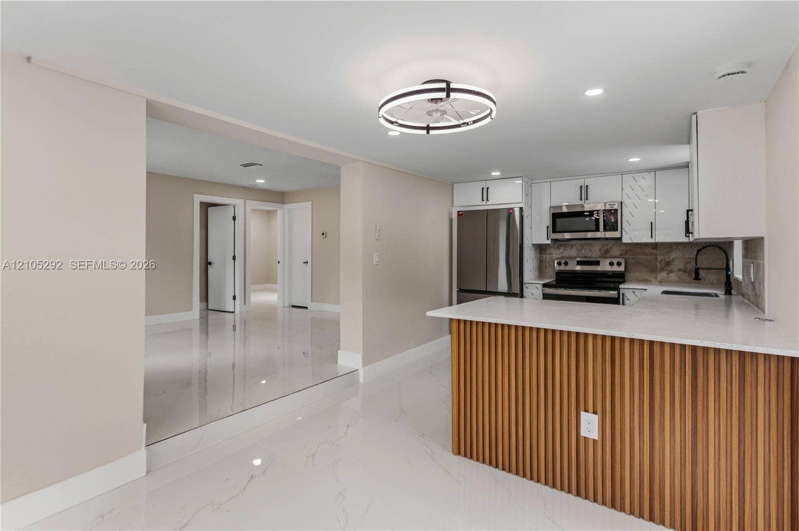 White tile flooring, kitchen island with slatted wood-finish paneling, white countertop, stainless steel appliances, and sleek white cabinetry