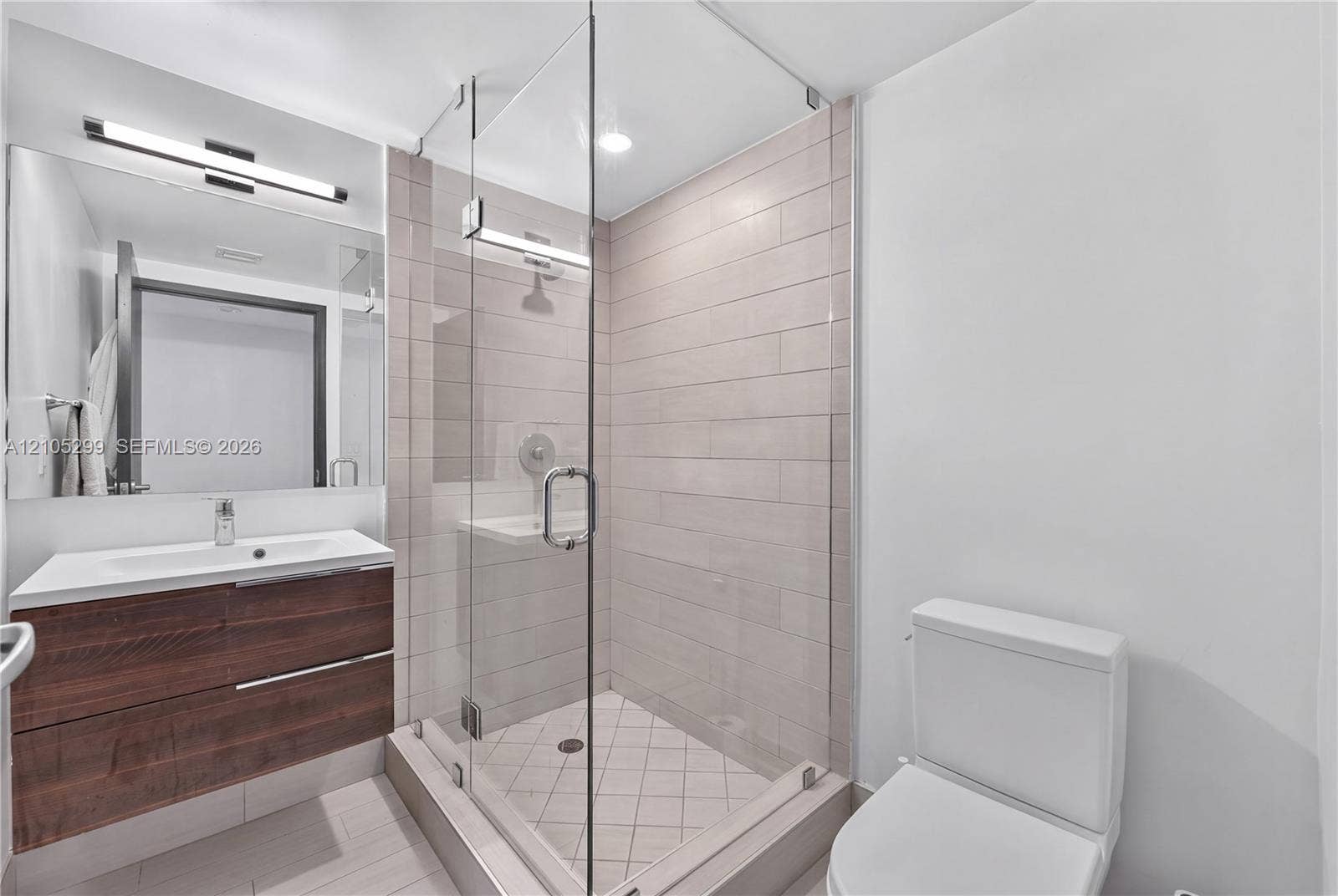Floating wood-finish vanity with an integrated sink, large vanity mirror, glass-enclosed shower with tile surround, and an elongated toilet