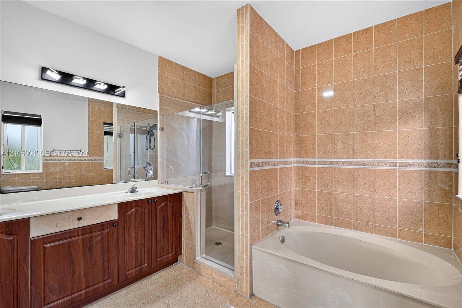 15217 SW 36th Terrace, Miami, FL 33185 Bathroom featuring a wood-finish vanity with a light-toned countertop, a soaking tub with tile surround, a glass-enclosed shower, a large wall-mounted mirror, and vanity lighting