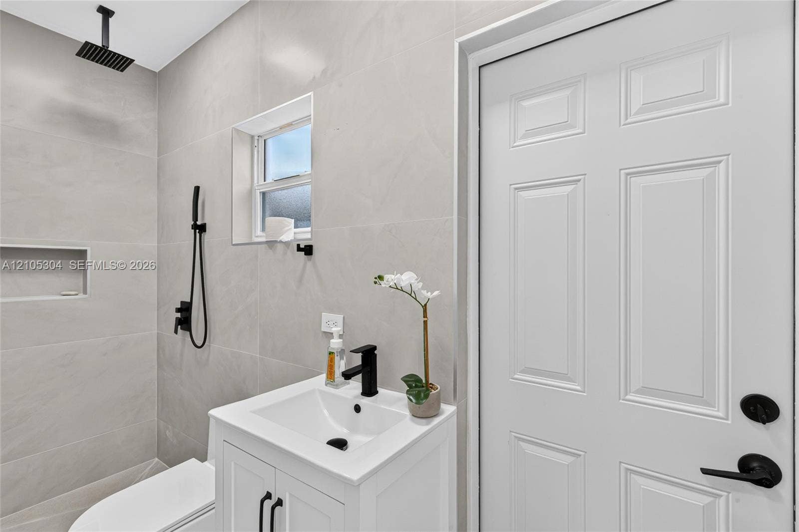 15217 SW 36th Terrace, Miami, FL 33185 Full-height neutral wall tiling, walk-in shower with rainfall showerhead and built-in wall niche, white vanity with rectangular basin and matte black fixtures, and six-panel interior door with matte black hardware