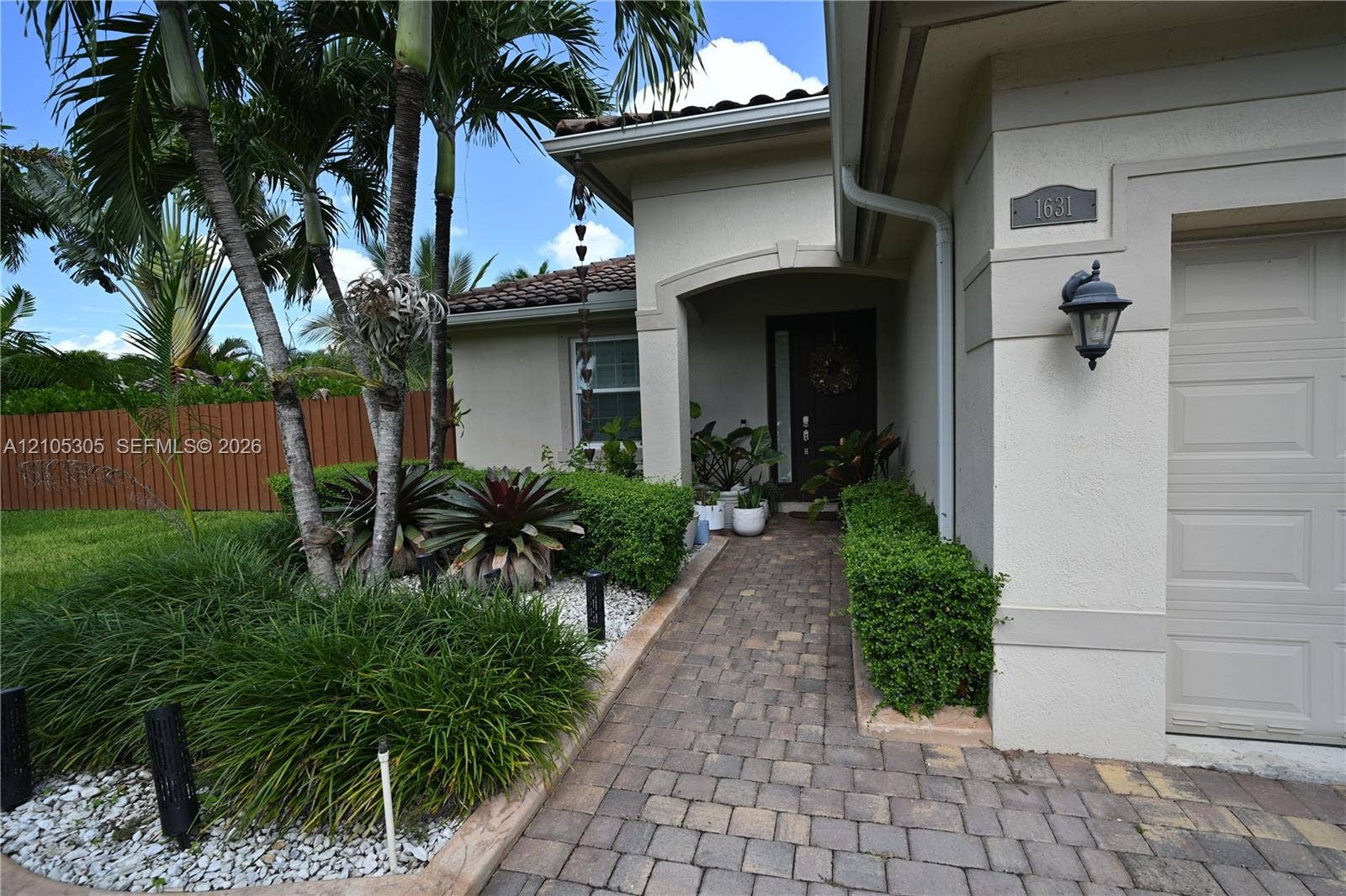Paver driveway and walkway, arched entryway, exterior wall sconce, and mature landscaping