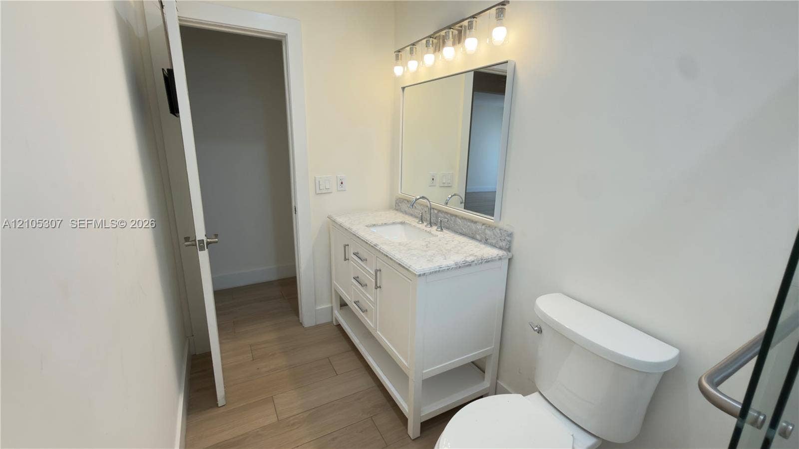 Bathroom vanity featuring stone-finish countertop, integrated sink, and white cabinetry, accompanied by a rectangular mirror, five-light fixture, and wood-finish flooring