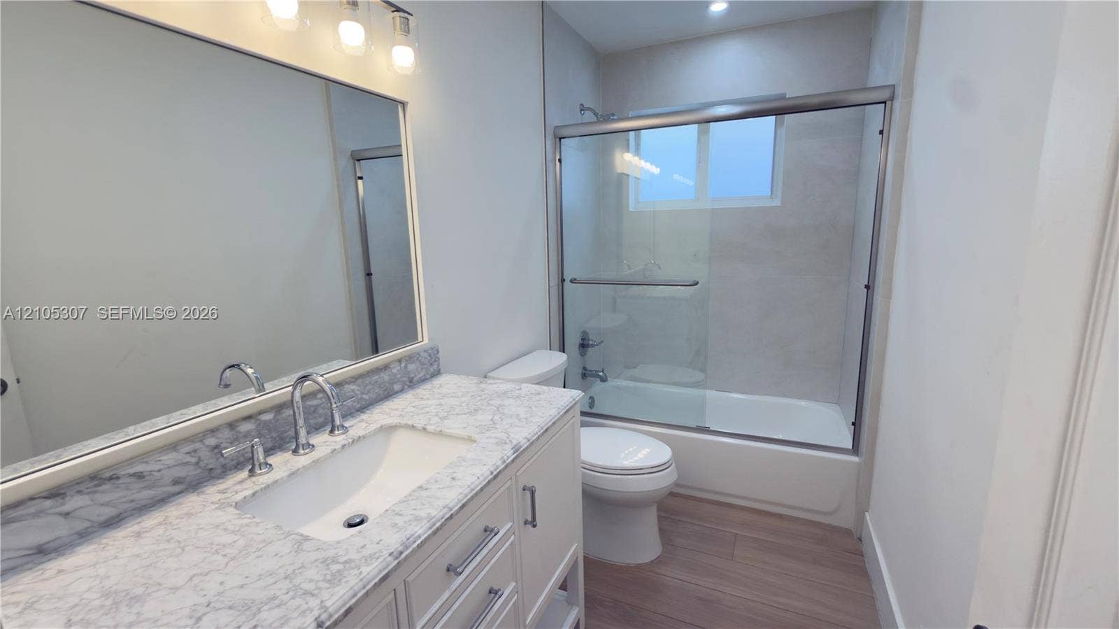 Bathroom with tub, eaturing a vanity with a stone-finish countertop, undermount sink, chrome faucet, large wall-mounted mirror with integrated lighting, and a tub-shower combination with a glass sliding enclosure