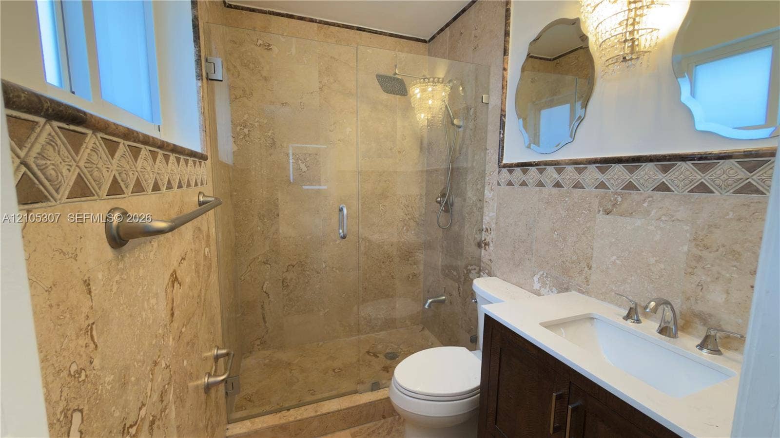 Bathroom featuring natural stone wall tile with decorative accent border, a glass-enclosed shower with a rainfall showerhead, and a vanity with a white rectangular sink and countertop