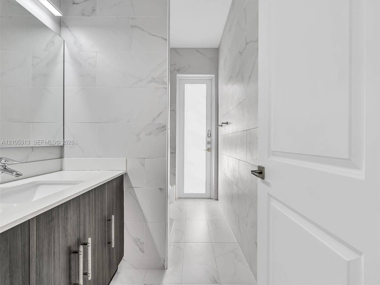 Bathroom featuring a wall-mounted mirror, vanity with a rectangular sink, wood-finish cabinetry, floor-to-ceiling tiled walls, and a glass-paneled door