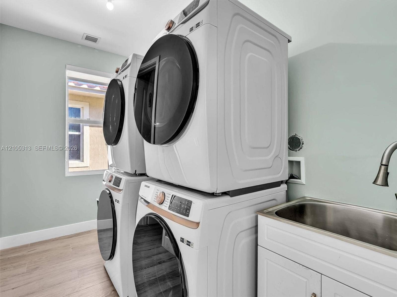 Laundry area featuring wood-finish flooring, stackable washer and dryer units, white cabinetry, a stainless steel basin, and a gooseneck faucet