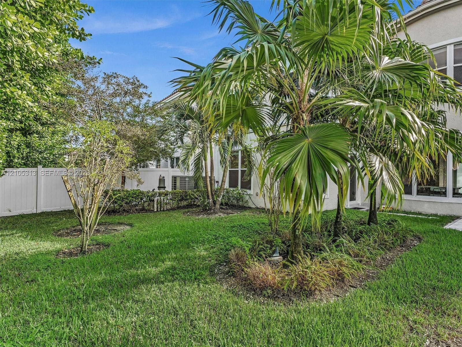 Backyard featuring a green lawn, mature palm trees, perimeter vinyl fencing, and structural siding exterior