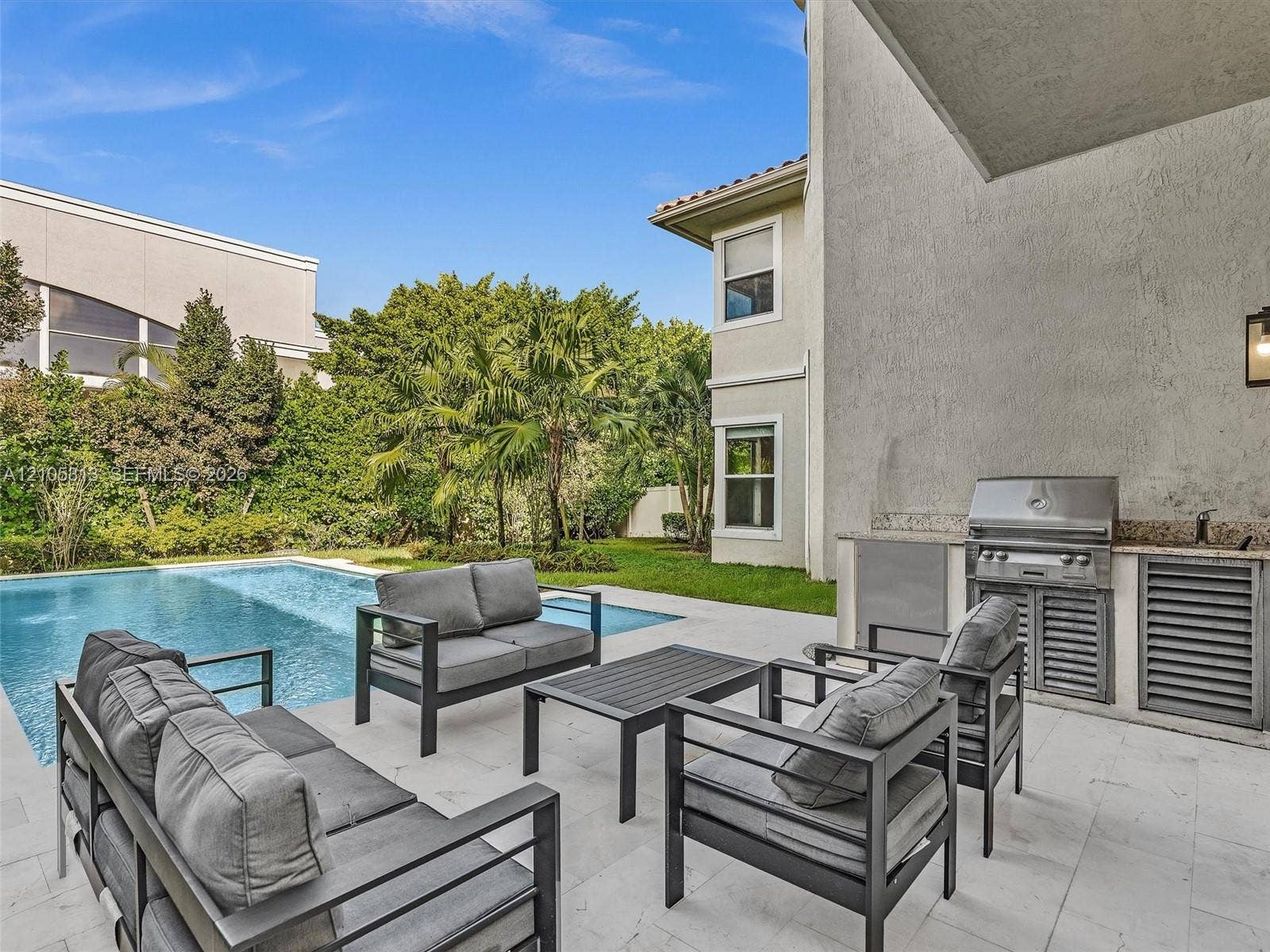 Outdoor living area featuring a covered patio with light-toned stone flooring, an integrated stainless steel built-in grill, stone countertop, swimming pool, and manicured landscaping