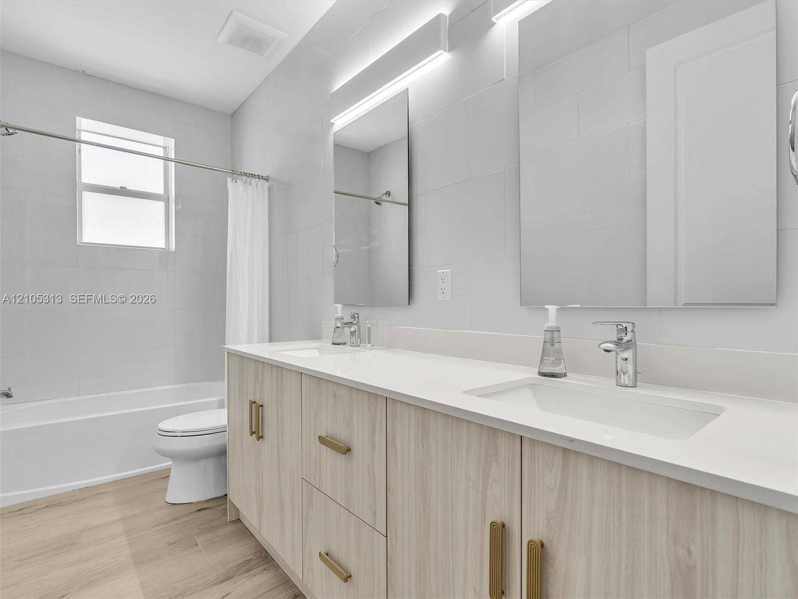 Dual-sink vanity with light wood-finish cabinetry, solid surface countertop, brass-tone hardware, and floor-to-ceiling wall tile