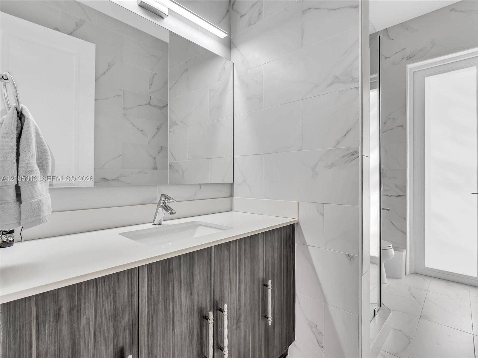 Vanity featuring dark wood-finish cabinetry, a rectangular undermount sink, and a solid-surface countertop, complemented by floor-to-ceiling stone-look wall tile and integrated strip lighting