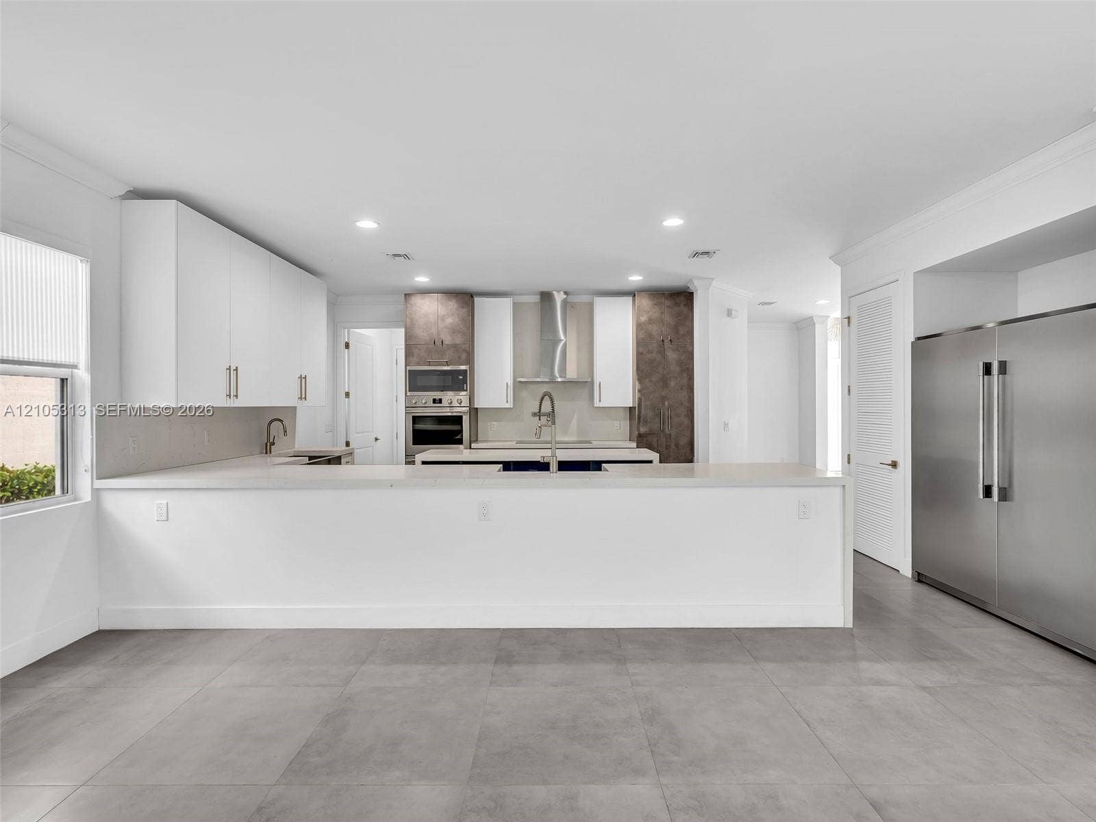 Kitchen featuring white flat-panel cabinetry, light-toned quartz countertops, stainless steel appliances, a center island with an undermount sink, and large-format gray tile flooring