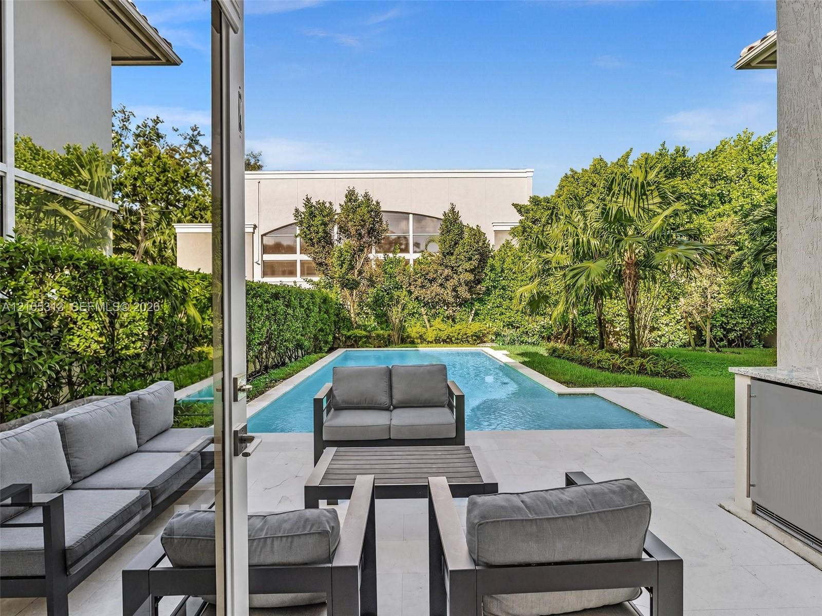 Rear patio featuring light-toned stone hardscaping, an outdoor kitchen with stone countertops, a rectangular swimming pool, and manicured landscaping with perimeter privacy hedges