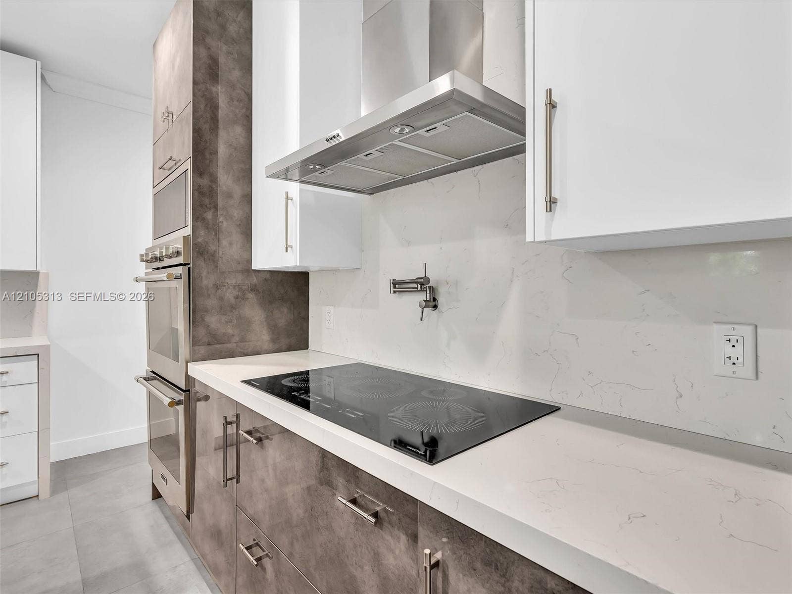 Kitchen featuring white flat-panel cabinetry, contrasting textured brown base cabinetry, a stone-patterned backsplash, a built-in stainless steel double oven, and a cooktop with a wall-mounted pot filler