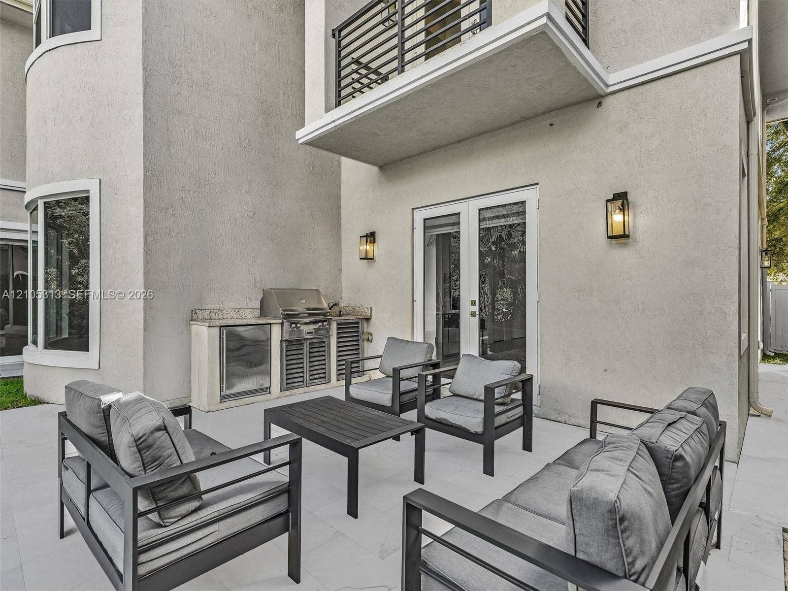 Patio space featuring stone flooring, a built-in outdoor kitchen with stainless steel grill and stone countertop, double glass doors, wall-mounted exterior lanterns, and a second-story balcony with metal railings