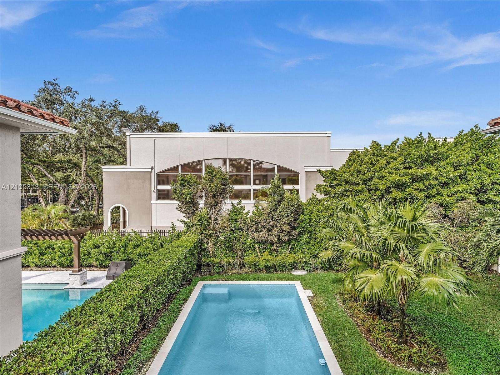 Rectangular swimming pool with light-colored tile border, manicured lawn, established shrubbery, and wood-framed pergola