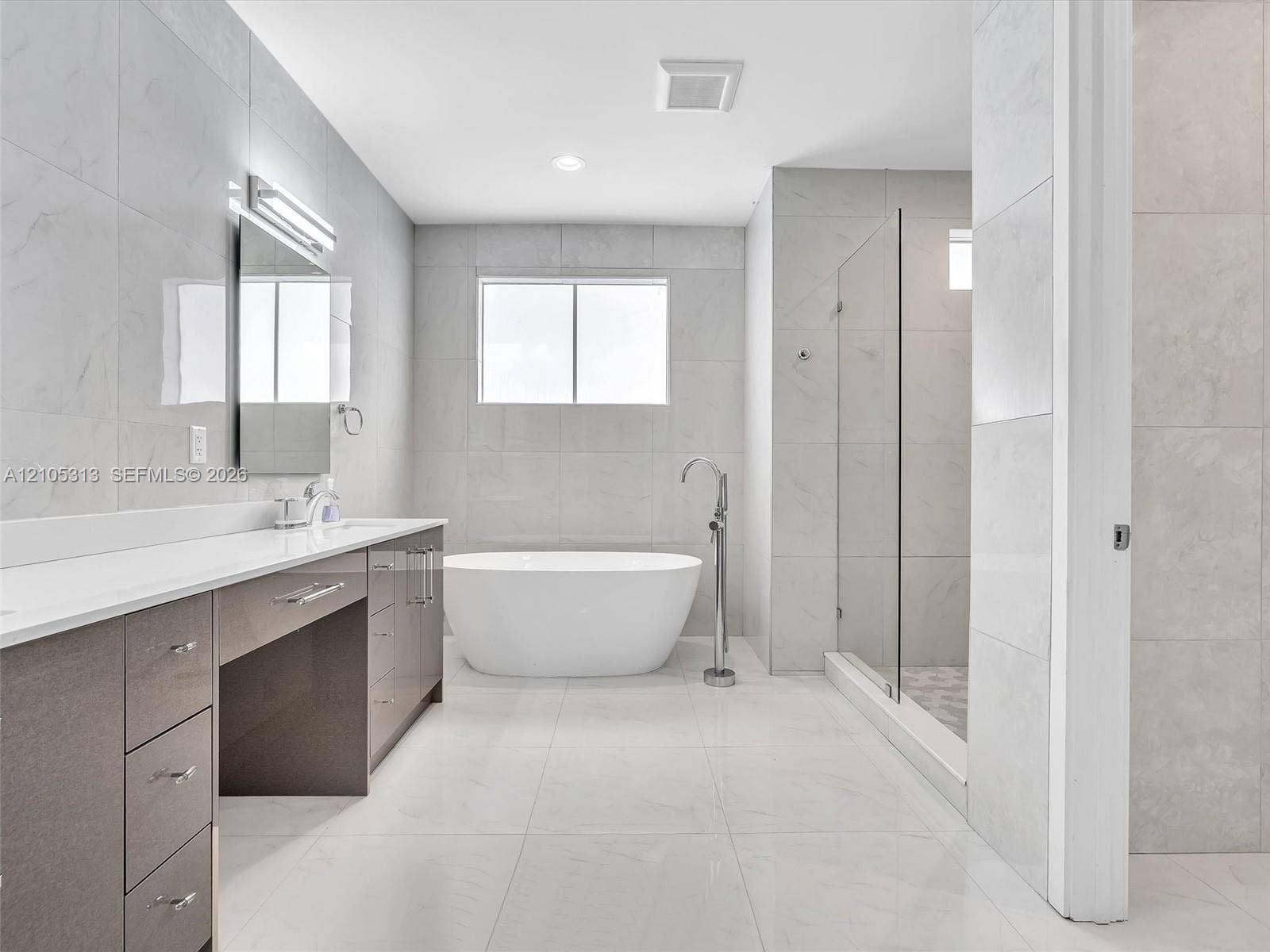 Dual-sink vanity with light-tone countertops, standalone soaking tub, walk-in glass-enclosed shower, and large-format tile flooring