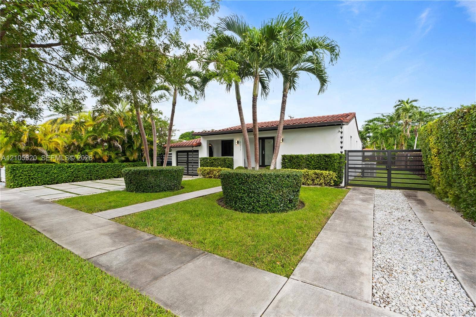 White exterior with a red tile roof, flanked by mature palm trees and lush hedges