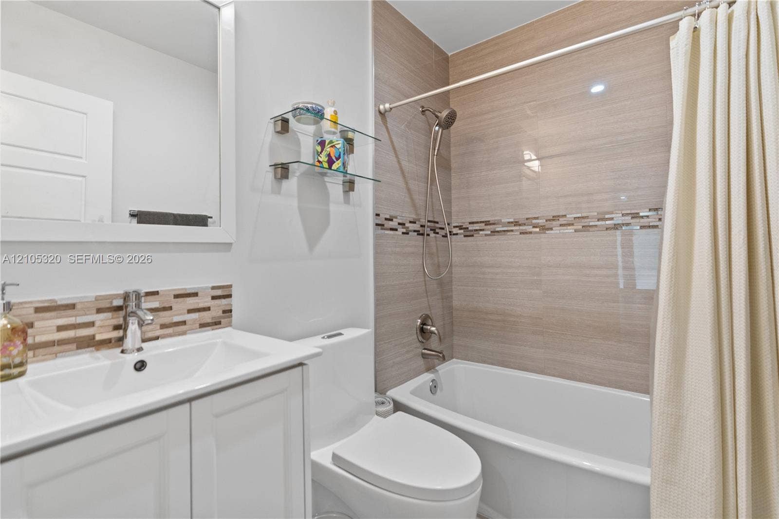 White vanity with integrated sink, chrome faucet, tiled backsplash, white toilet, and a tiled shower/tub combination with a decorative accent band