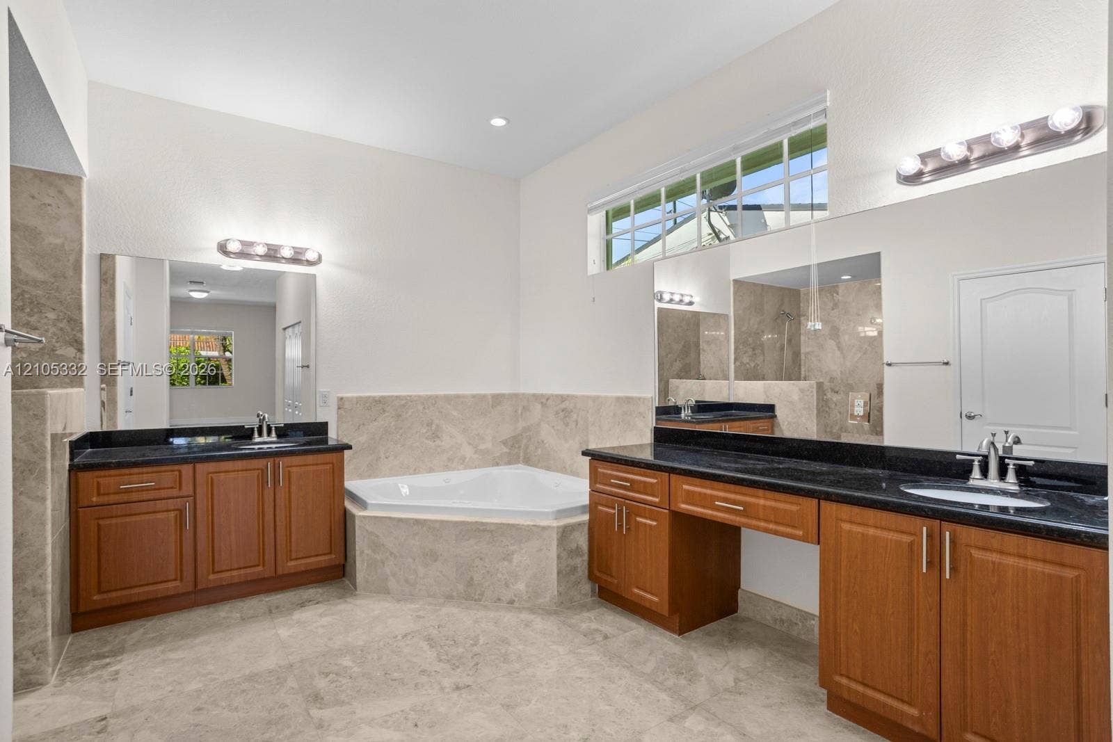 Dual vanity with wood cabinetry and dark countertops, corner soaking tub, large wall-mounted mirrors, and transom windows