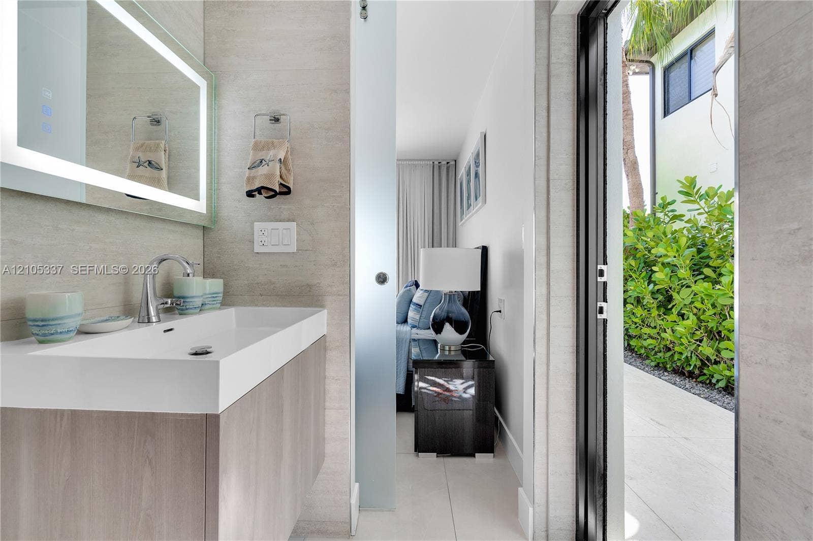 3322 NE 166th Street, North Miami Beach, FL 33160 Modern floating vanity with a rectangular integrated sink, LED-backlit mirror, frosted glass sliding door, and stone-finish wall tile