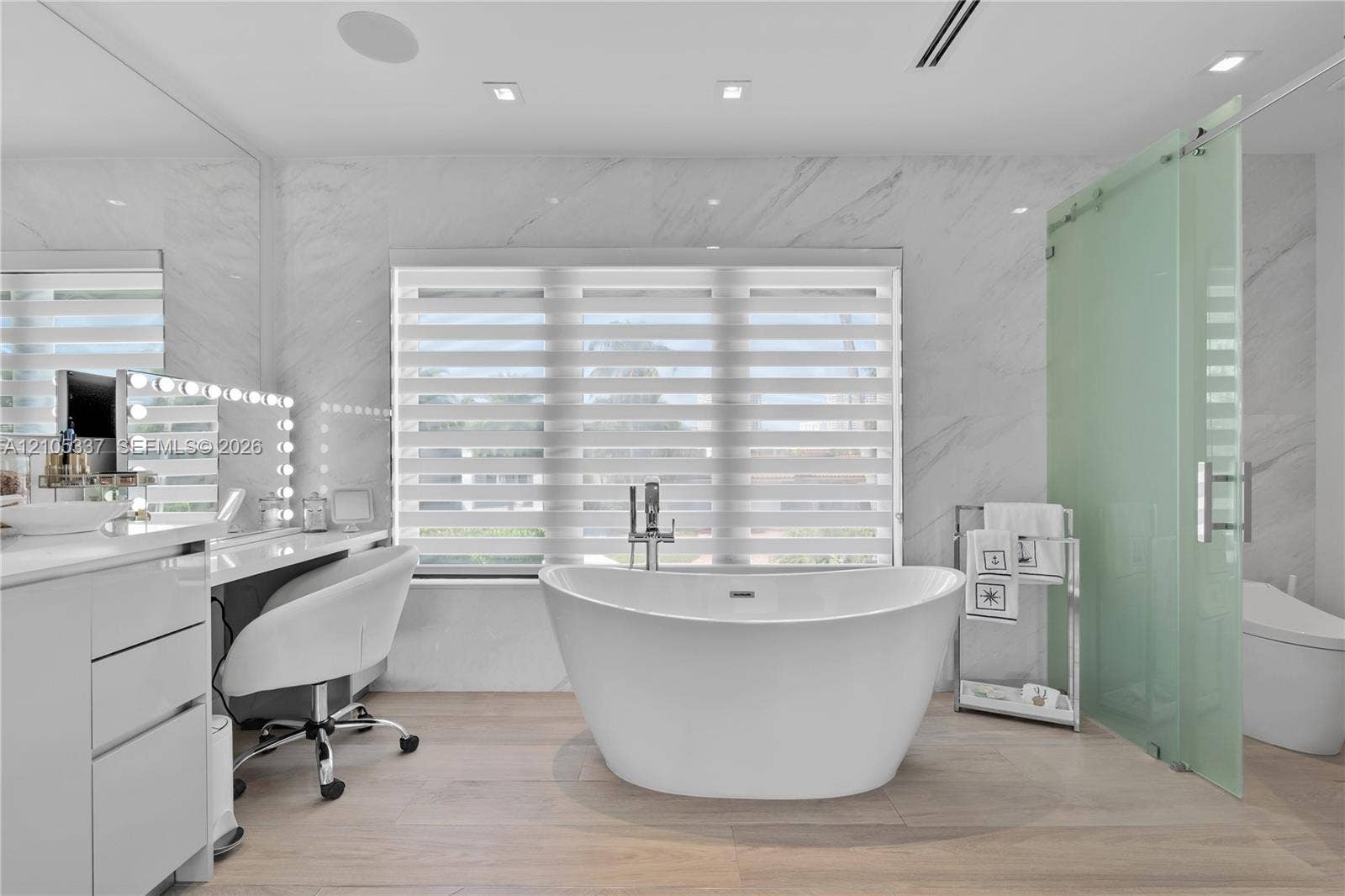 3322 NE 166th Street, North Miami Beach, FL 33160 Freestanding soaking tub with a chrome floor-mounted faucet, wall-to-wall stone surfacing, wood-finish flooring, and a frosted glass-enclosed shower