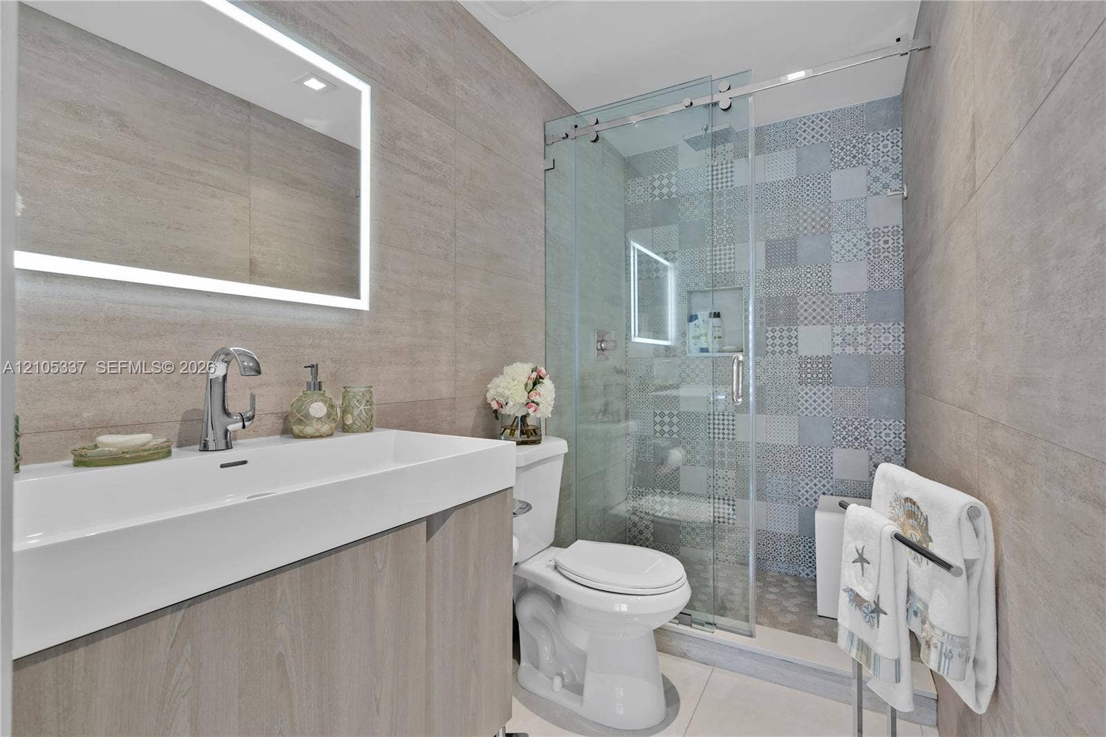 3322 NE 166th Street, North Miami Beach, FL 33160 Wall-mounted rectangular basin with a single-handle faucet, integrated LED-backlit mirror, wood-finish vanity cabinetry, glass-enclosed shower with decorative patterned tile wall, and large-format wall tiling