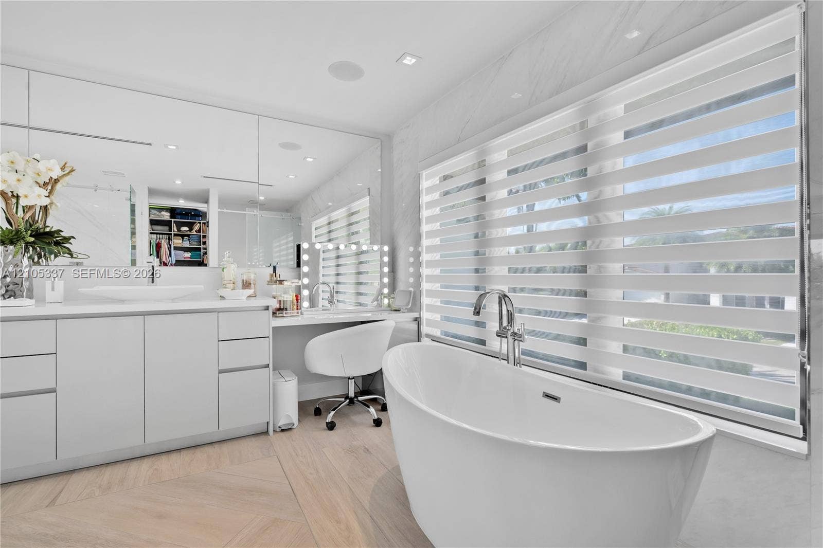 3322 NE 166th Street, North Miami Beach, FL 33160 Freestanding soaking tub with a chrome floor-mounted faucet, custom vanity with integrated sink and stone-finish countertop, full-wall mirror, and wood-finish flooring in a herringbone pattern