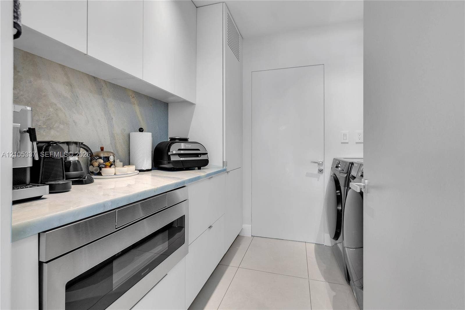 3322 NE 166th Street, North Miami Beach, FL 33160 Utility room featuring flat-panel white cabinetry, stone-surface countertop, full-height stone backsplash, built-in stainless steel microwave, and large-format floor tile