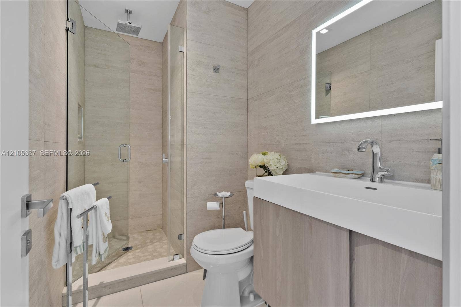 3322 NE 166th Street, North Miami Beach, FL 33160 Bathroom featuring large-format neutral wall tile, walk-in glass shower with integrated niche, floating vanity with vessel-style sink, and LED-backlit mirror