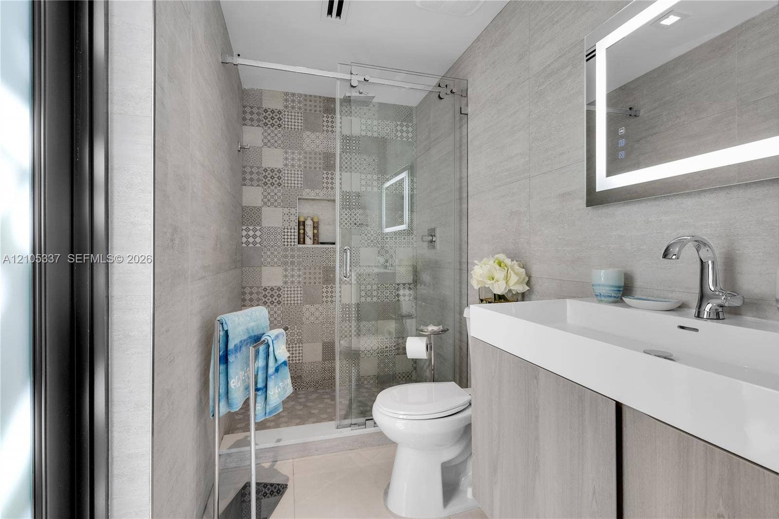 3322 NE 166th Street, North Miami Beach, FL 33160 Modern bathroom featuring a wood-finish vanity with an integrated basin, LED-backlit mirror, and a walk-in shower with patterned tile accents, glass enclosure, and a built-in recessed shelf