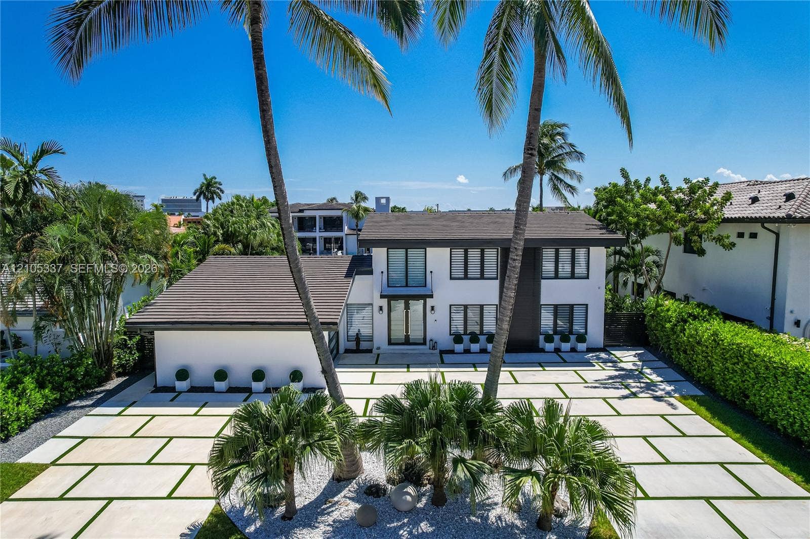 3322 NE 166th Street, North Miami Beach, FL 33160 Two-story residence featuring a crisp white exterior, dark-toned roof, paved driveway with grass grid detail, multi-pane windows, and a central balcony