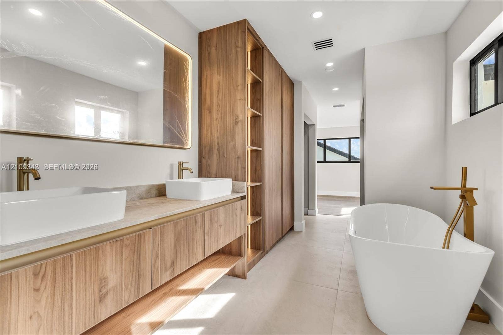 Floating wood-finish vanity with dual vessel sinks, custom integrated cabinetry featuring open shelving, brass-tone fixtures, and a freestanding soaking tub