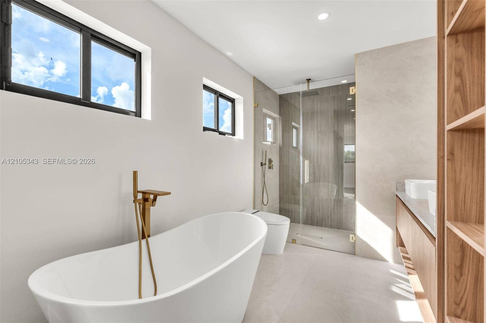 Freestanding soaking tub with a brass floor-mounted faucet, walk-in glass-enclosed shower, wall-mounted dual-flush toilet, floating wood-finish vanity with countertop sinks, and built-in wood shelving