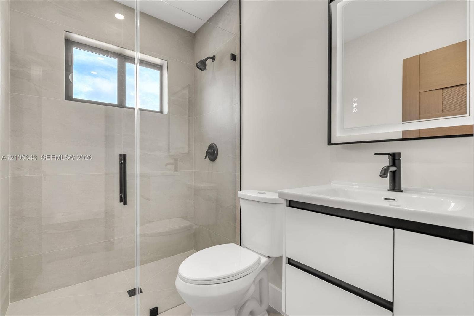 Modern bathroom featuring a frameless glass-enclosed shower, matte black plumbing fixtures, a white vanity with integrated basin, and a backlit wall-mounted mirror