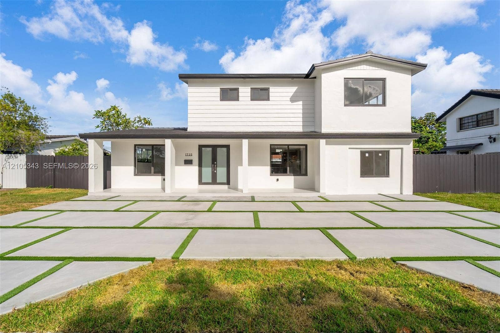 Two-story residence featuring a white exterior, covered front porch, double glass-panel entry doors, and geometric concrete paver driveway with integrated turf strips