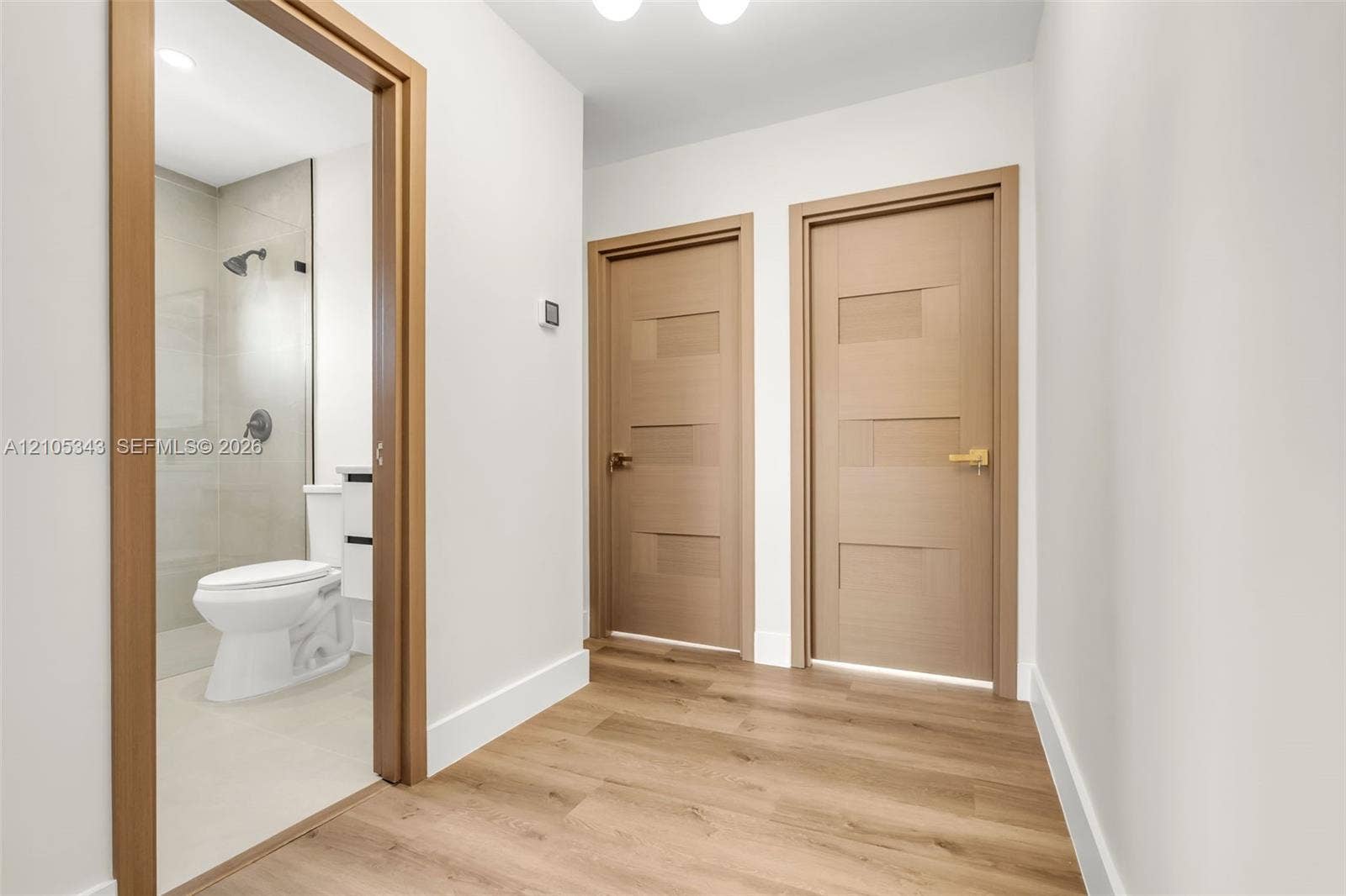 Hallway featuring wood-finish flooring, two wood-grain interior doors with geometric paneling, and a secondary doorway revealing a bathroom with a glass-enclosed shower and neutral tile finishes