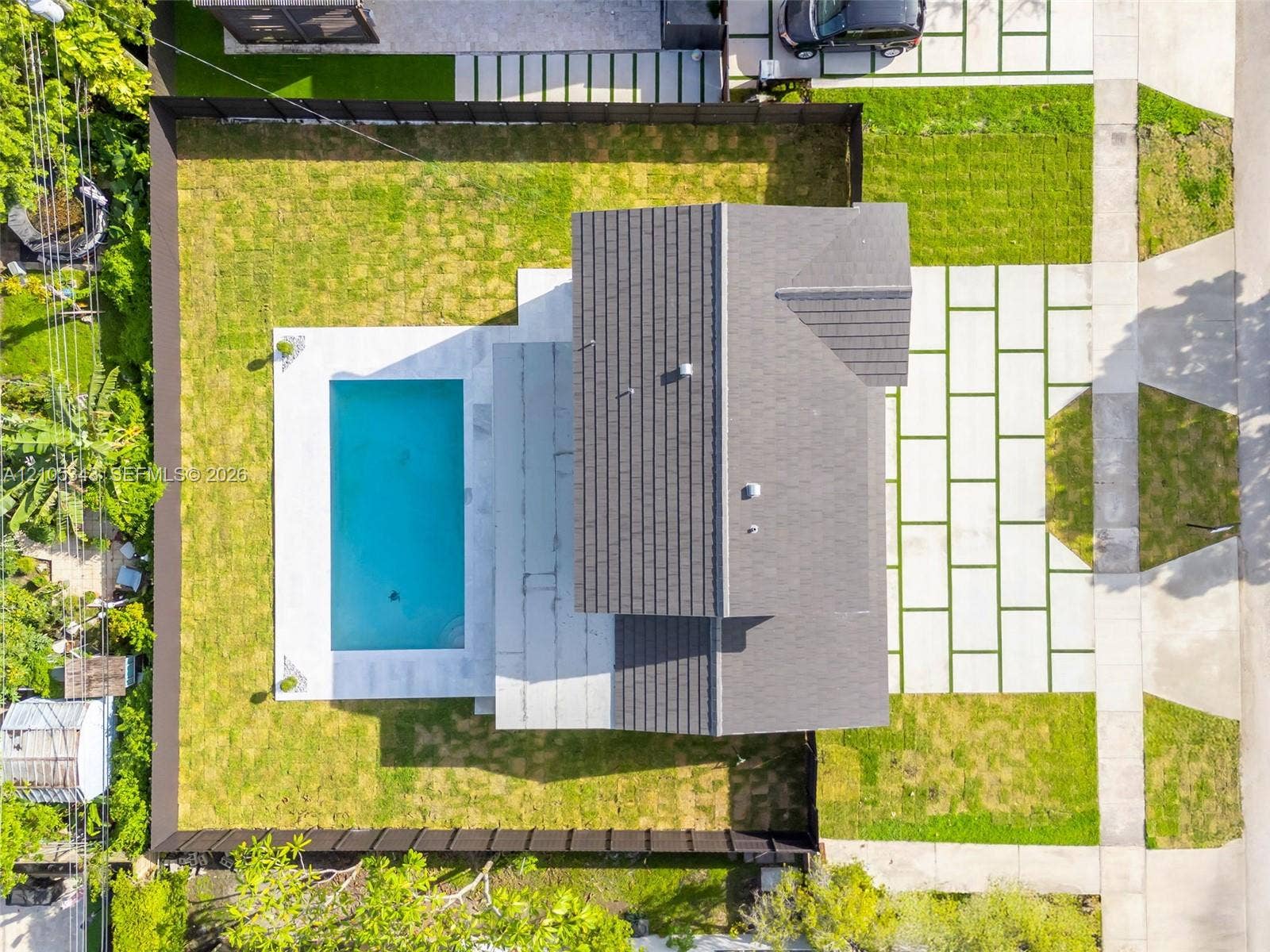 Aerial perspective featuring a rectangular swimming pool, concrete paver driveway with integrated turf ribbons, fenced perimeter, and shingled roof