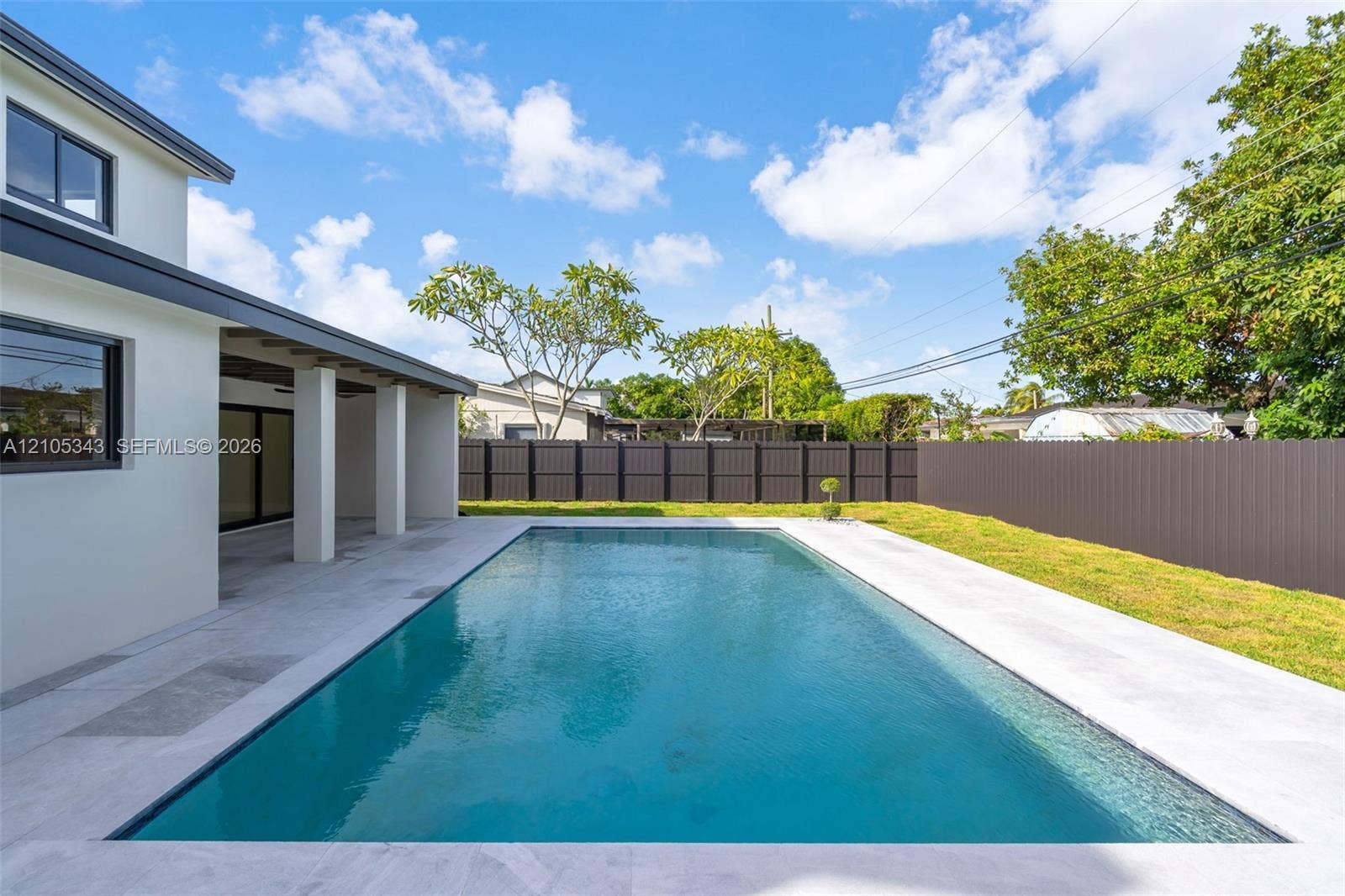 Rectangular swimming pool featuring a light-colored concrete deck, covered patio, and perimeter privacy fencing