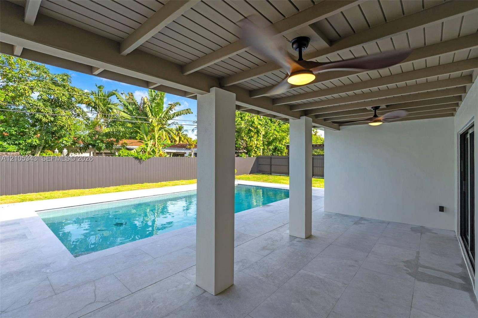 Covered patio featuring large-format stone flooring, a timber-beam ceiling, and integrated ceiling fans, overlooking an in-ground swimming pool enclosed by a privacy fence