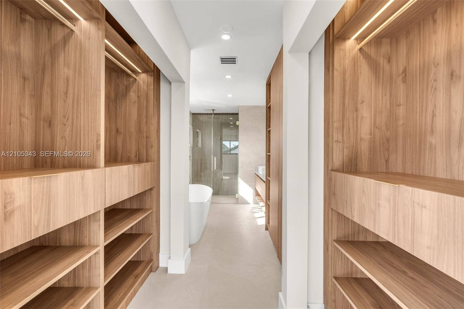 Custom wood-finish cabinetry with integrated lighting, open shelving, freestanding soaking tub, glass-enclosed shower, and large-format tile flooring