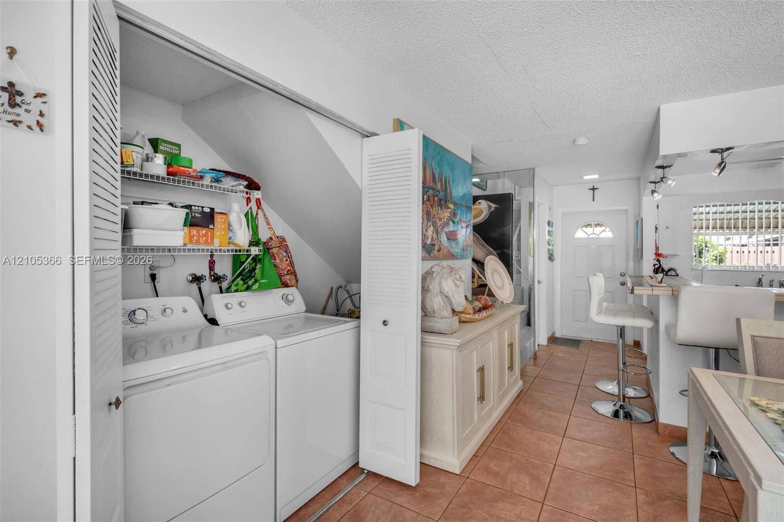 Built-in laundry closet with wire shelving, tiled flooring, front entry door with arched window, and track lighting