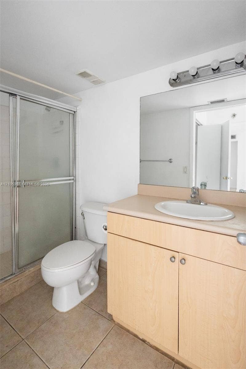 9066 W Atlantic Boulevard, #412, Coral Springs, FL 33071 Bathroom featuring light wood-finish cabinetry, a neutral countertop with an integrated oval basin, ceramic floor tiling, a walk-in shower with a glass enclosure, and a wall-mounted four-bulb vanity fixture