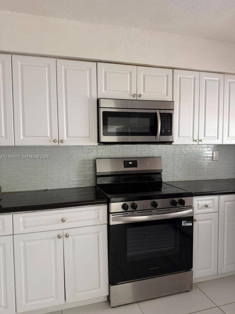 White shaker-style cabinetry, black countertops, stainless steel range, coordinating stainless steel microwave, and a tiled backsplash