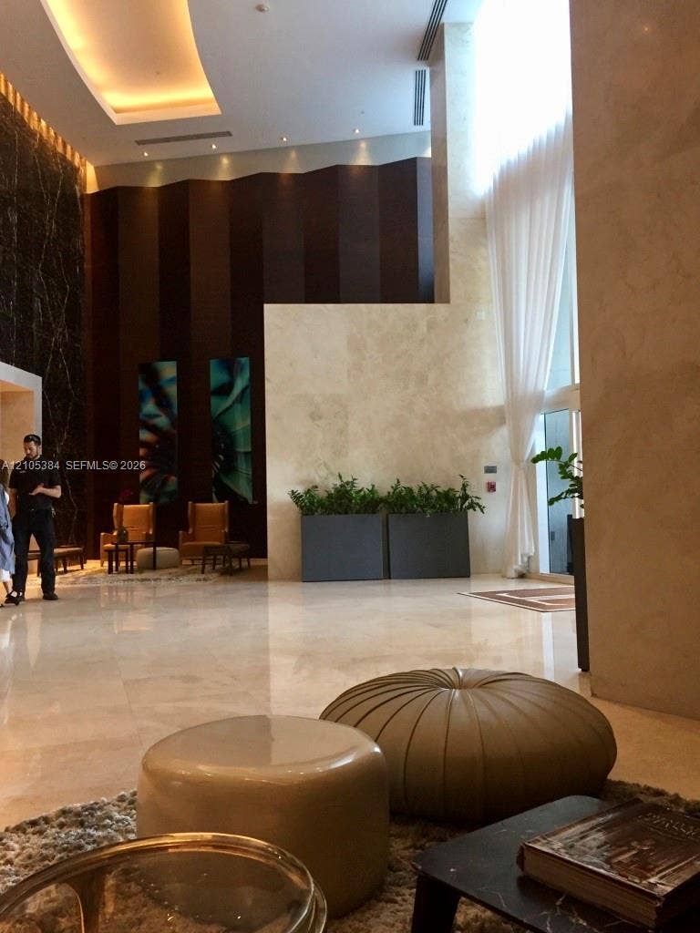 16001 Collins Avenue, #2803, Sunny Isles Beach, Sunny Isles Beach, FL 33160 High ceilings with recessed lighting, full-height window with sheer drapes, dark wood-paneled accent wall, light-toned stone flooring, and built-in planters