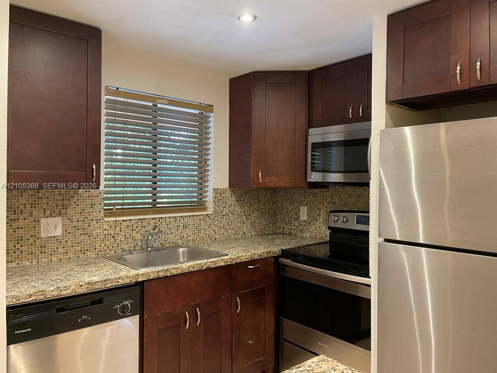 Kitchen with wood cabinetry, stainless steel appliances, granite-style countertops, mosaic tile backsplash, and a stainless steel sink