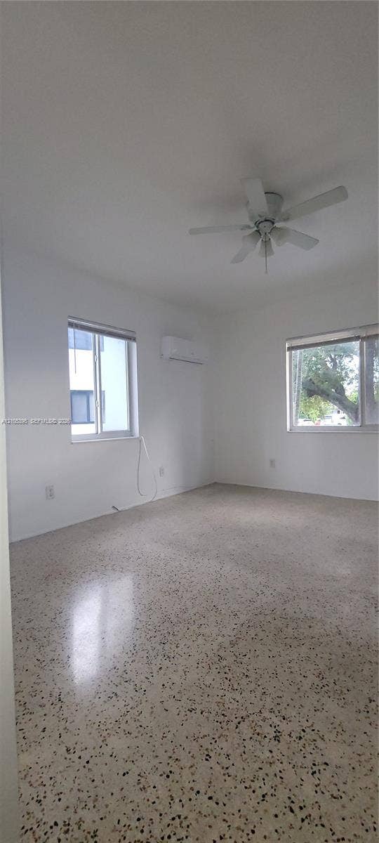 Spacious room featuring terrazzo flooring, white walls, multiple windows, a ceiling fan, and a wall-mounted air conditioning unit