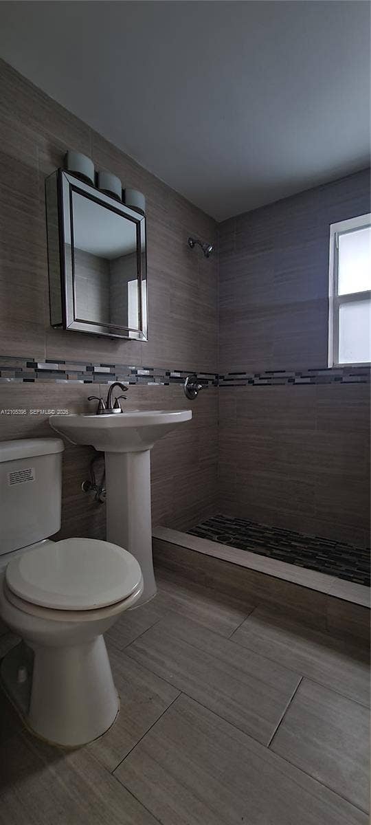 Pedestal sink with chrome faucet, built-in medicine cabinet with integrated lighting, tiled shower with textured floor, full wall tiling with decorative accent band, and a window for natural light