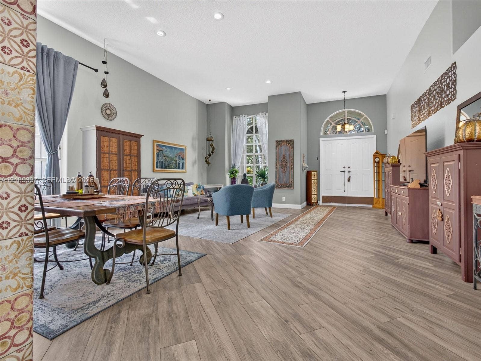 High-ceiling living and dining area featuring wood-finish flooring, recessed lighting, double-entry doors, and expansive windows
